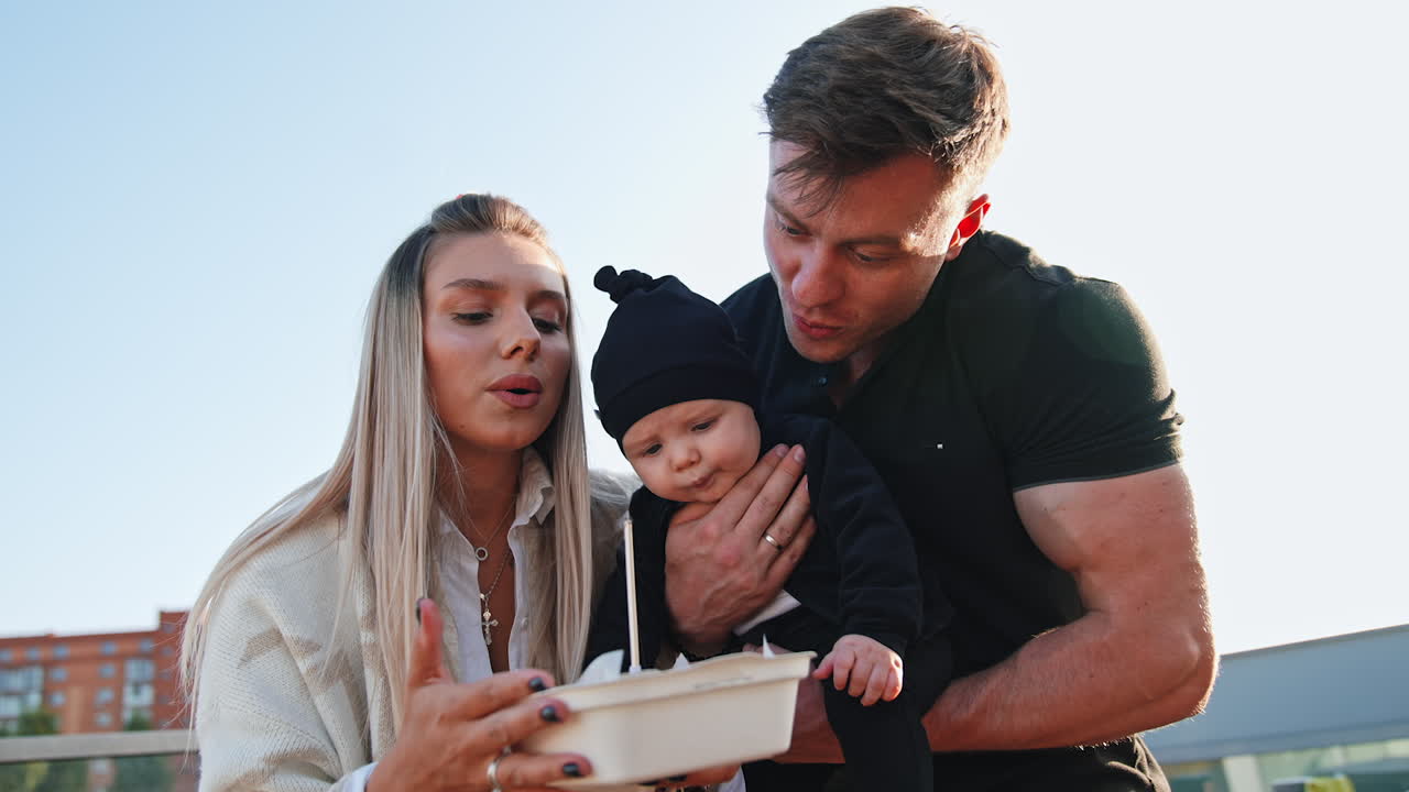 Caucasian couple with a newborn blowing a candle on the cake. Happy family celebration outdoors. Low angle view.