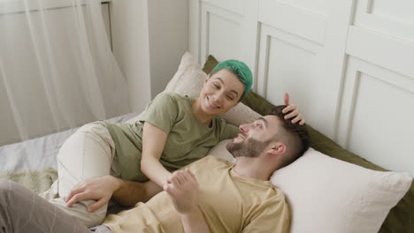 mujer sonriente acariciando el cabello de su novio y hablando con él mientras están acostados juntos en la cama