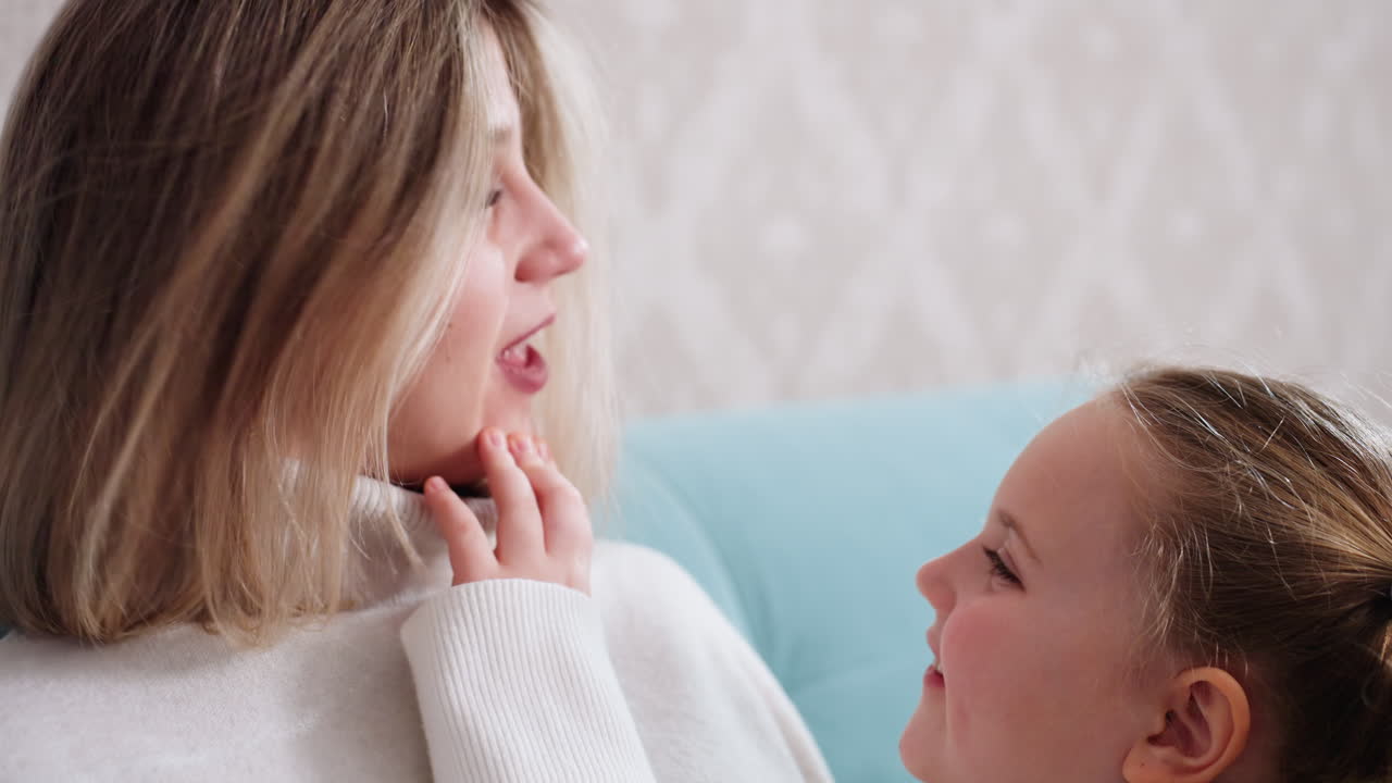 Smiling mother sitting on sofa wearing cozy white sweater while young daughter touches her chin affectionately, sharing warm family bonding moment filled with love, joy, tenderness