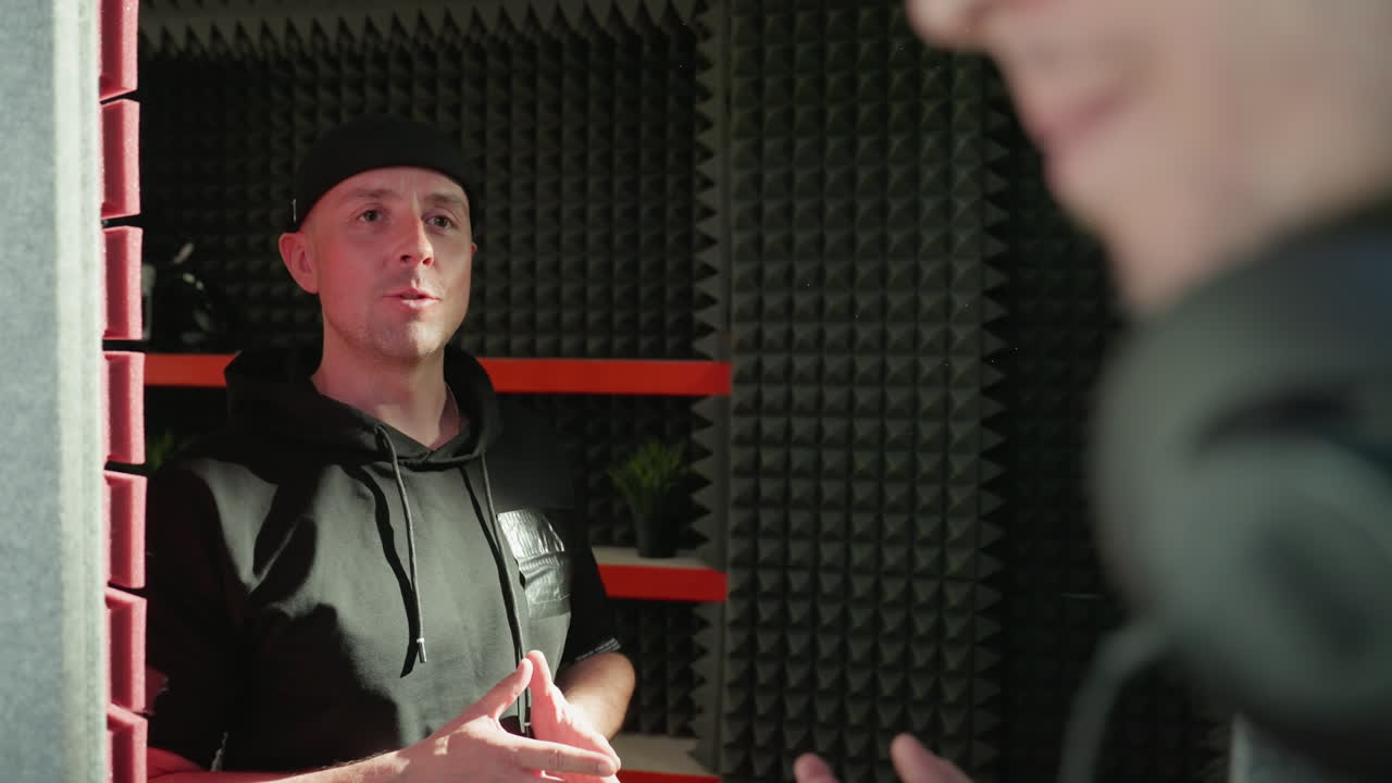 검은 옷을 입은 남자가 은색과 검은색의 스튜디오 배경에 표시적인 손짓으로 활기찬 대화를 나누고 있습니다.