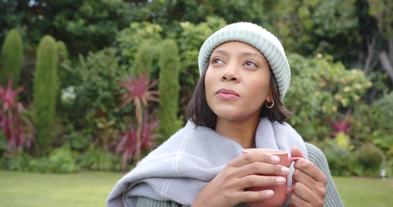 Donna afroamericana seduta in giardino, avvolta in una coperta, che tiene in mano una tazza e alza lo sguardo dall'aria fresca.