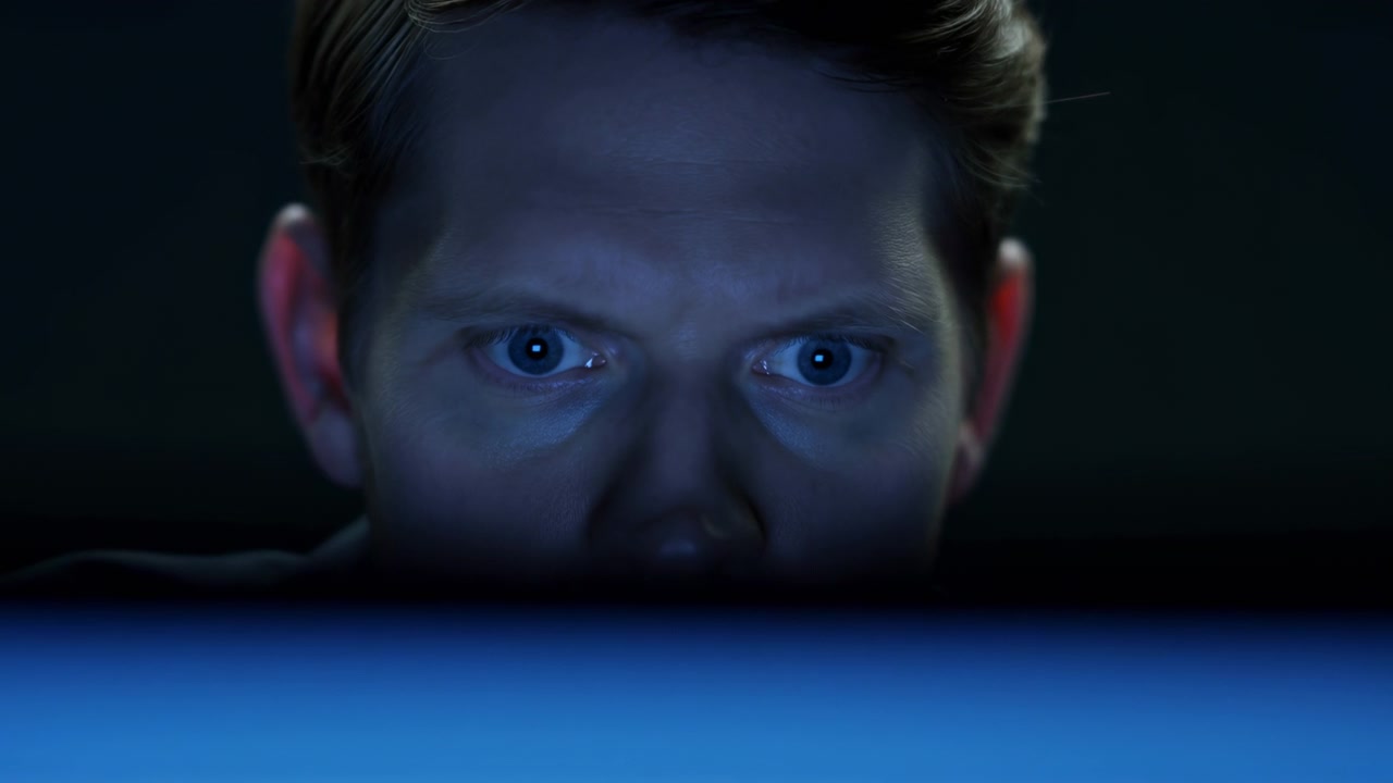 Close frontal view shows a man staring steadily toward a glowing monitor, with cool blue illumination across his face, dark background, centered framing, and minimal visible movement.