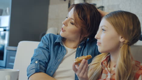 Portrait-shot-of-the-pretty-woman-and-her-blonde-teen-daughter-sitting,-talking-and-resting-while-a-girl-eating-bread-with-peanut-butter.-Close-up.-Indoors