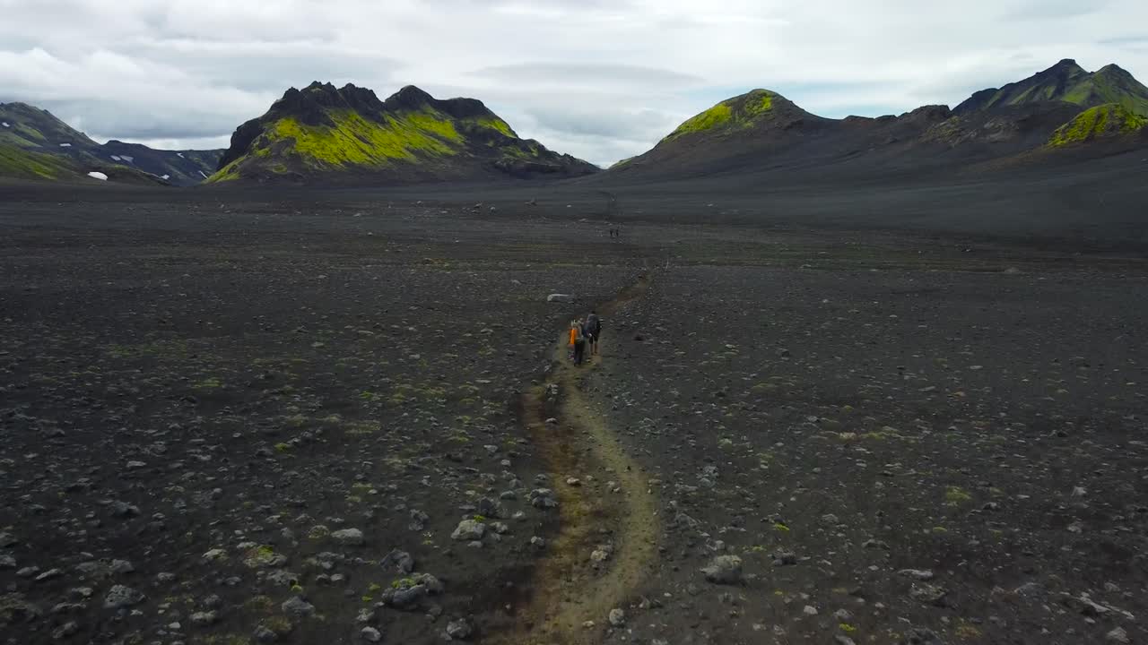 Aerial drone flying behind two hikers walking on dark soil and rocky Iceland landscapes where large mountains with green moss and snow are visible in the background during a cloudy day. low to ground.