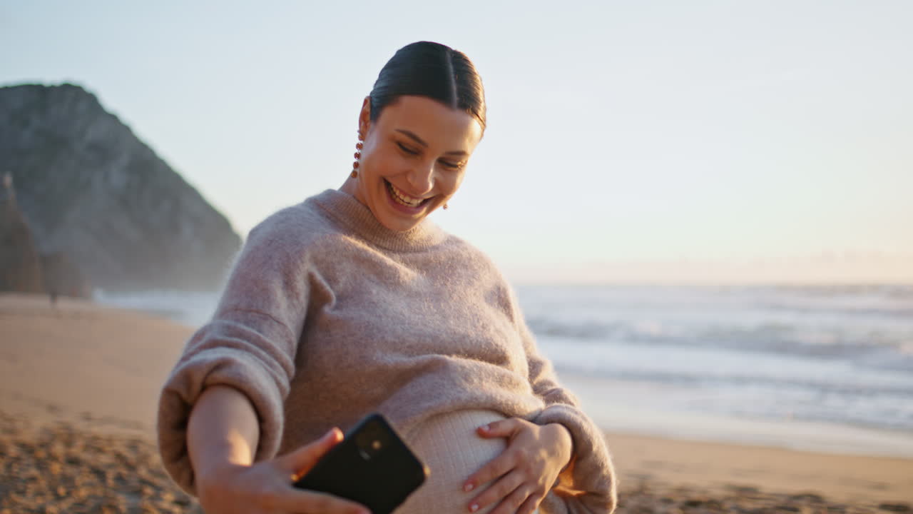 임신한 여성이 스마트폰으로 비디오 블로그를 작성하고 샌디 코스트에서 가까이 걸어가고 있습니다.