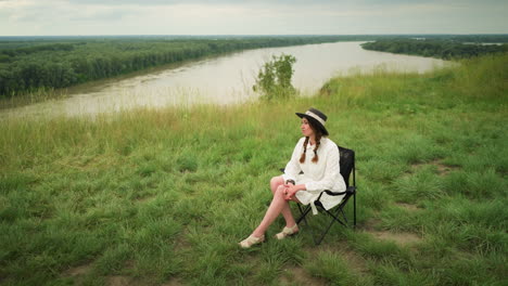 an elegant woman wearing a stylish hat and a white dress is seated on a chair in a lush grass field beside a tranquil lake. she poses gracefully, capturing a serene moment under a cloudy sky