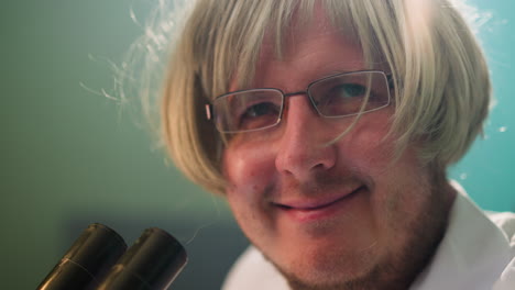 a facial view of a smiling scientist wearing a blonde wig and glasses, sitting in a laboratory with a microscope with light reflection seen on his head