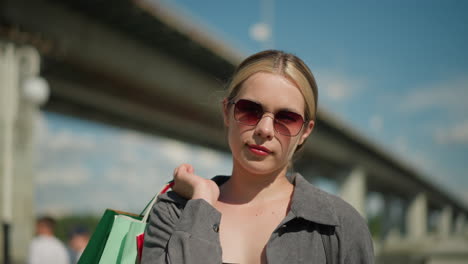 white woman wearing grey shirt and sunglasses carries shopping bag backward with a focused expression, she stands outdoors with a blurred background, and two people are seen passing by behind her