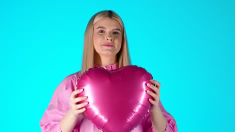 una mujer rubia bonita sosteniendo un globo de corazón púrpura y luego lo lanza, fondo azul de estudio