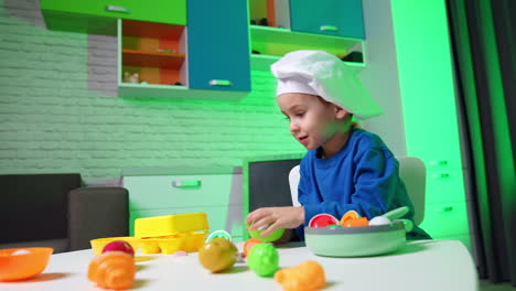 lovely smiling baby boy in white cap fits in the chair. kid plays with plastic toy crockery and food.
