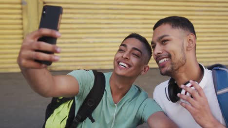 dos felices amigos varones de raza mixta sentados, tomando selfies en la calle
