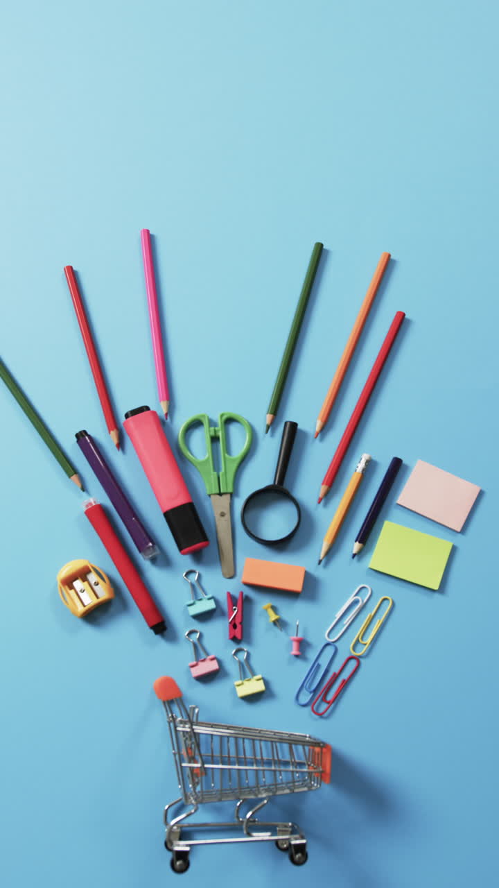 Vertical video of overhead view of shopping trolley and school items on blue background