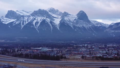 Lkw-Mit-18-Rädern-Und-Autos-Fahren-Auf-Der-Autobahn-Im-Vordergrund-Mit-Der-Bergkette-Der-Drei-Schwestern-In-Canmore-Alberta-In-Kanada,-Mitreißende-Dynamische-Drohnenaufnahme