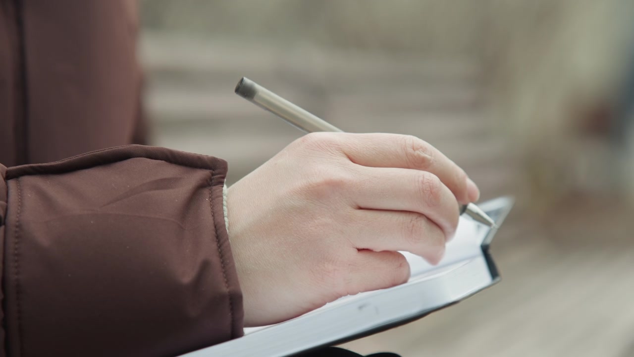 Closeup hands flipping pages of planner on bench, brown coat, pen poised, organized layout, purposeful posture urban planner scheduling goals, neat handwriting, overcast park background, steady