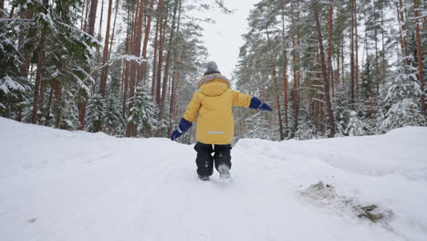 A-little-boy-of-3-4-years-old-runs-in-the-winter-forest-a-view-from-the-back-in-slow-motion-in-a-yellow-jacket.-The-concept-of-winter-fun-and-active-recreation-freedom-and-a-happy-childhood