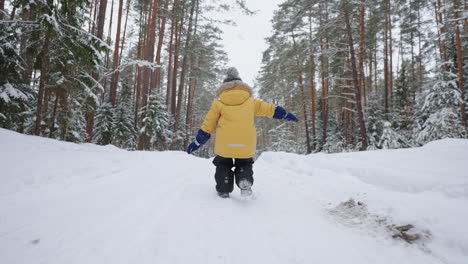 un niño pequeño de 3-4 años corre en el bosque de invierno una vista desde atrás en cámara lenta en una chaqueta amarilla. el concepto de diversión de invierno y recreación activa libertad y una infancia feliz