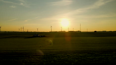 Volando-Lejos-Del-Parque-Eólico-Sobre-Campos-Verdes-Al-Atardecer,-Silueta-De-Aerogeneradores-Lejos-En-El-Fondo,-Puck,-Pomorskie,-Polonia,-Dron-Aéreo