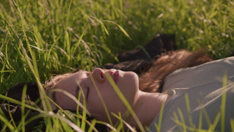 close-up of young woman lying in a sunlit grassy field, lost in thought, surrounded by tall green grass that gently frames her face, warm glow of sunlight in the background