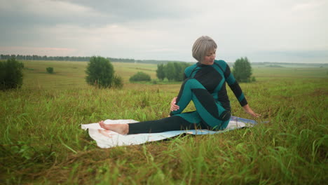 old woman seated on yoga mat in half seated twist practicing yoga, closing her eyes with head slightly raised, in a vast grassy field with trees lined up in the distance under a cloudy sky