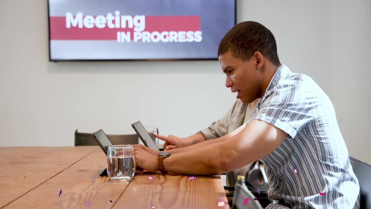 Two colleagues leaning tapping tablets when confetti falls, showing phone during business meeting