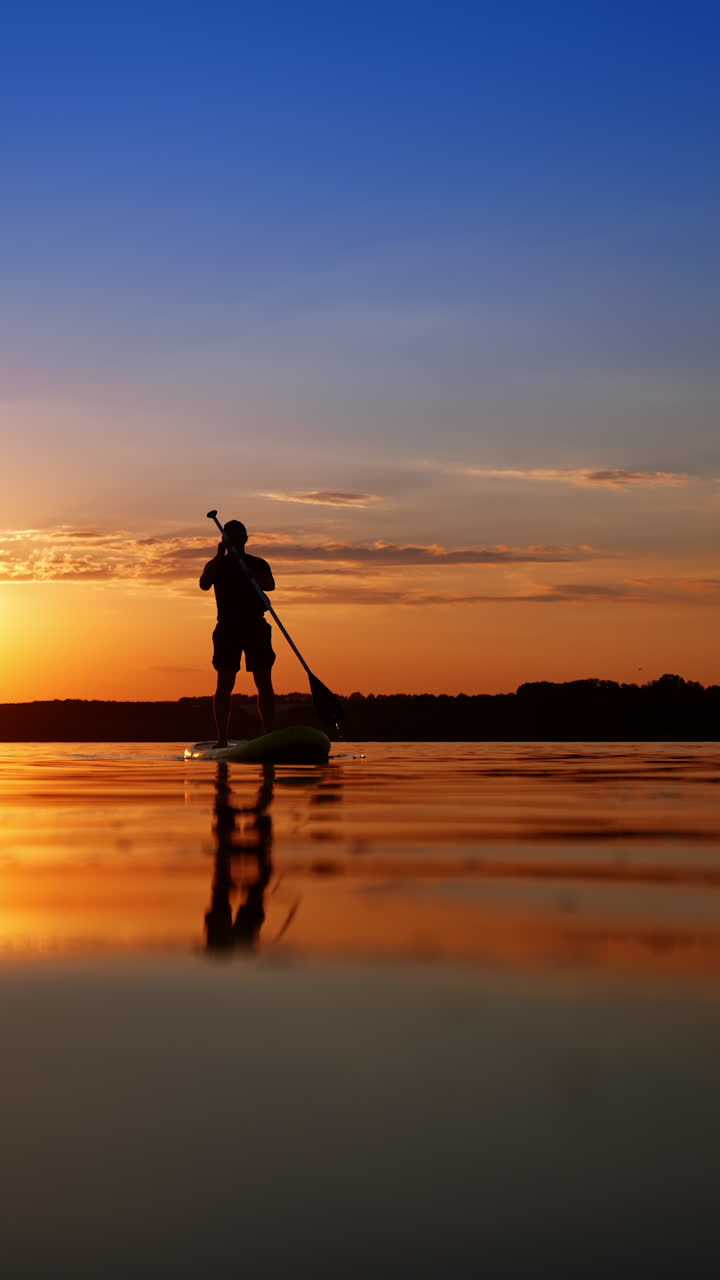 Male silhouette wearing a cap approaches floating on the sup board. Man uses an oar to move by the water at sunset. Low angle view. Vertical video.