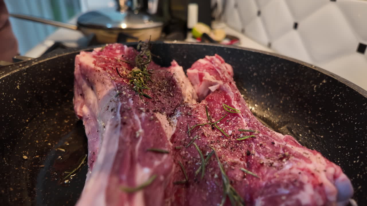 Adding salt on a big red beef stake on a black pan with rosemary ready for cooking, black pepper on top