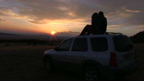 a silhouette of a couple sitting on top of a suv at sunset