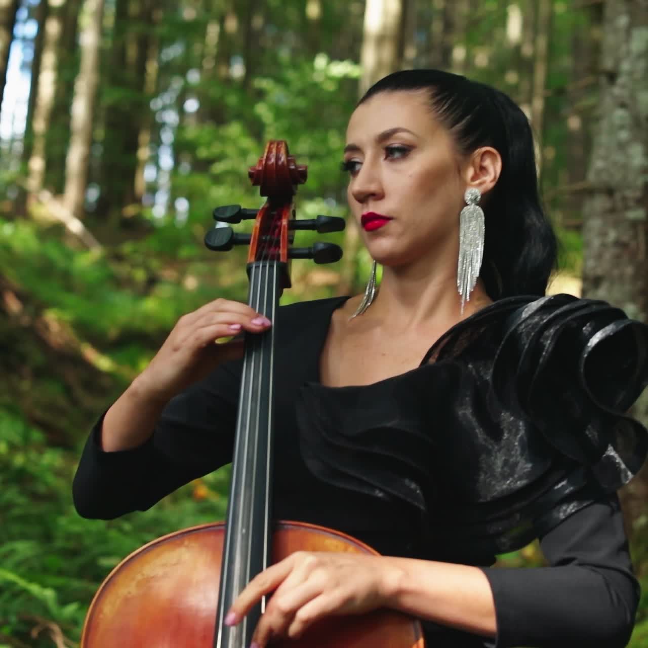 Attractive woman with musical instrument. Beautiful musician in black dress performing music in the forest. Pretty lady playing the cello outdoors.
