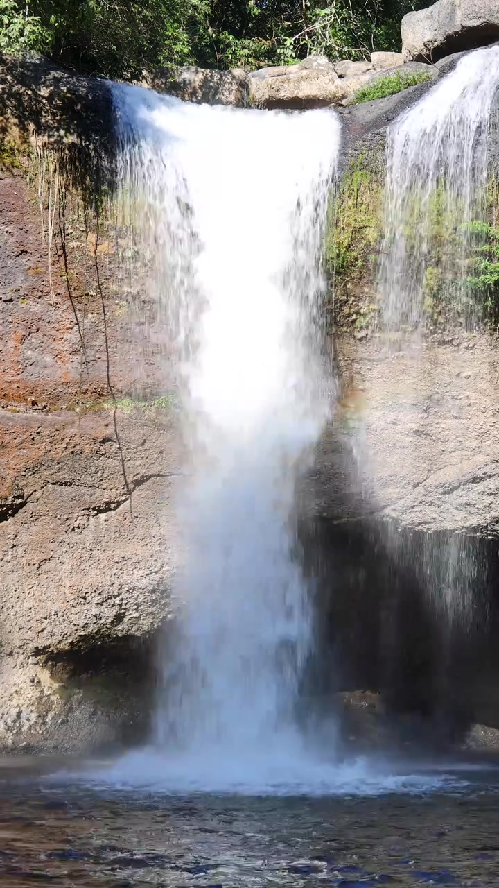 Una cascata si getta da una scogliera rocciosa in una pozza cristallina, circondata da una foresta lussureggiante alla luce del giorno.