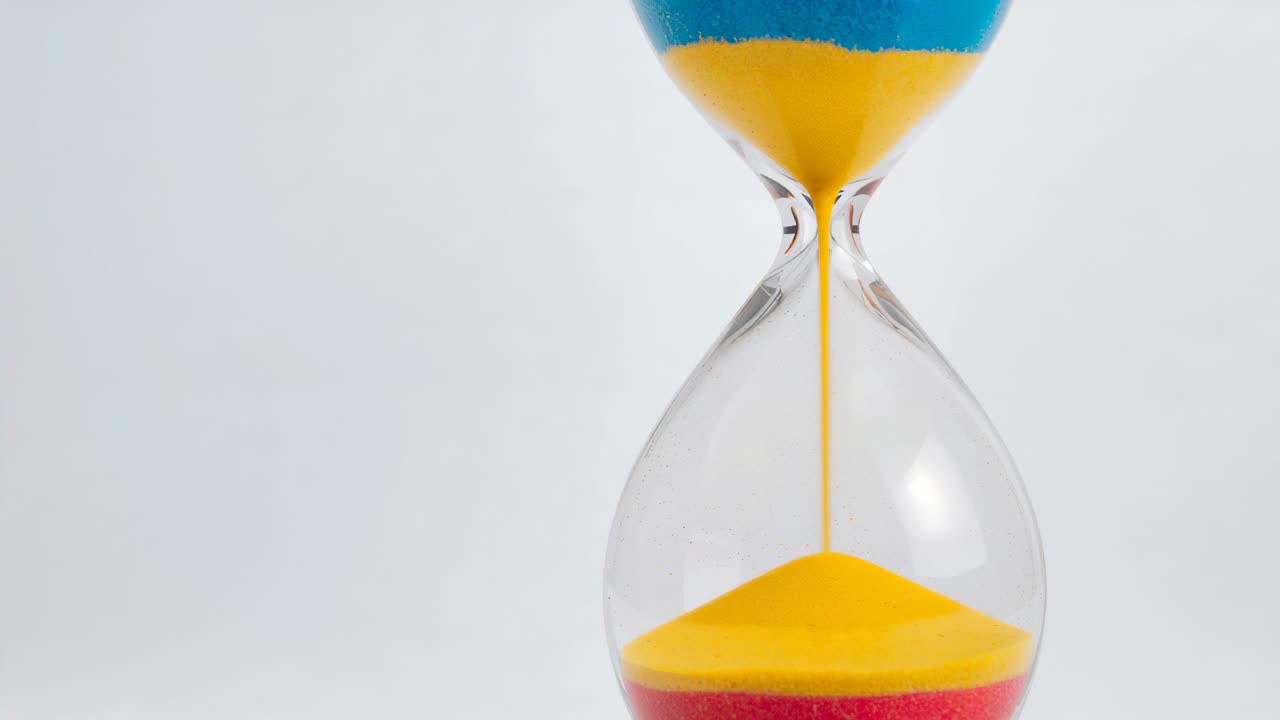 Starting glass hourglass trickling yellow sand through narrow tube in studio with blue and red sand