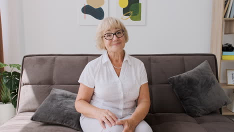 Happy-Senior-Woman-Sitting-On-Sofa-In-Living-Room-Looking-At-Camera-And-Greeting-While-Having-A-Video-Call