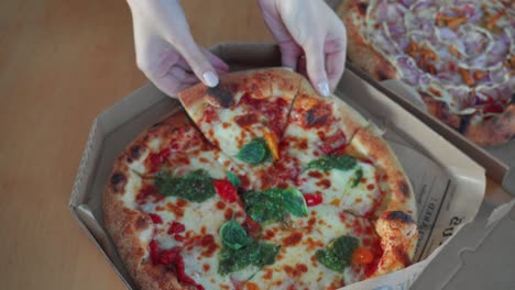 top down shot of a hand taking a slice of tasty neapolitan pizza with pesto sauce in a cardboard pizza box on the table