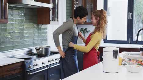 feliz pareja de lesbianas caucásicas poniendo delantales y abrazándose en la cocina soleada