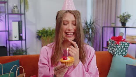 happy young woman celebrating birthday party, makes wish blowing burning candle on small cupcake