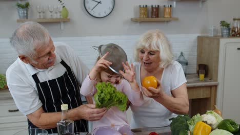 Senior woman and man with grandchild girl making a funny dance with strainer and vegetables at home