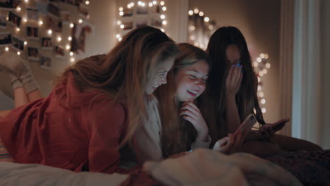 happy teenage girls lying on bed using smartphone watching online entertainment laughing having fun sleep over on weekend