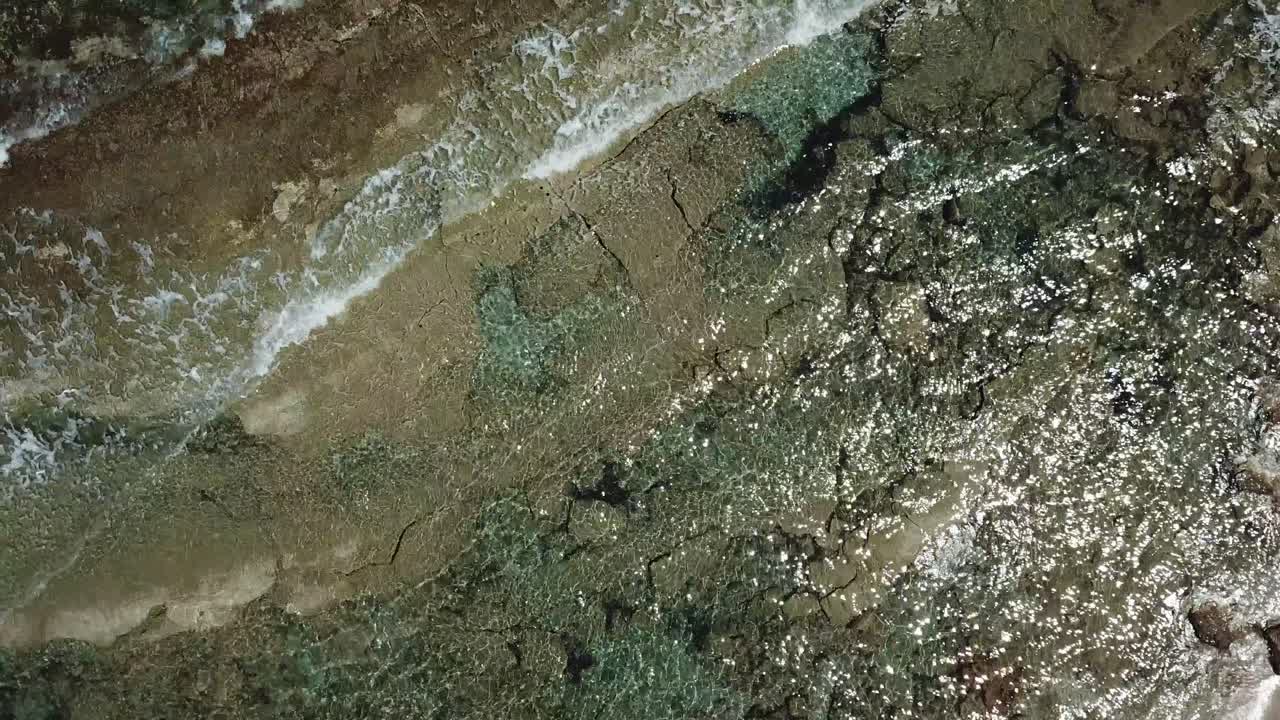 Aerial drone shot of tranquil sea waves softly lapping against a rocky shoreline in Crete, Greece, during a peaceful and sunny day with clear skies and serene coastal scenery