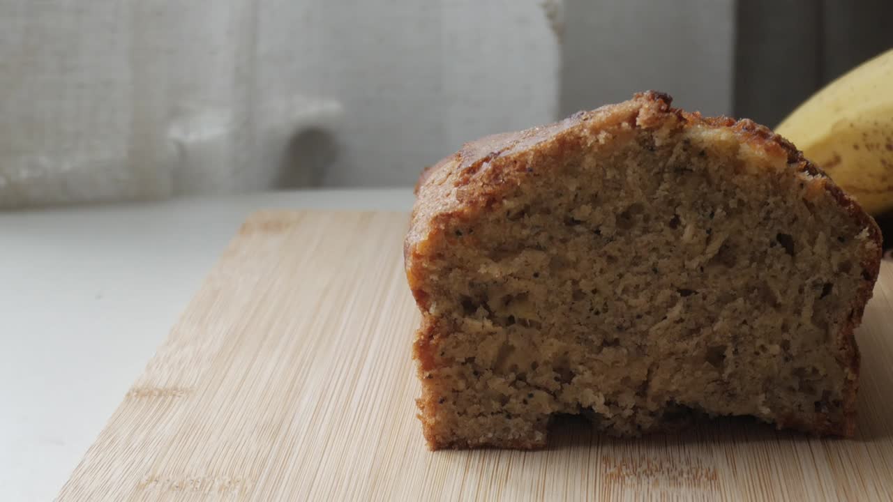 vista derecha del pan de plátano cortado por la mitad en una tabla de madera con un plátano en la parte posterior.