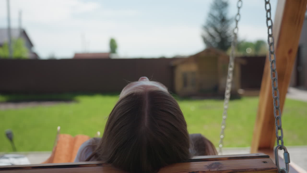 Rear view of woman with long brown hair sitting quietly on wooden swing bench, gazing at bright green lawn and wooden shed on sunny day