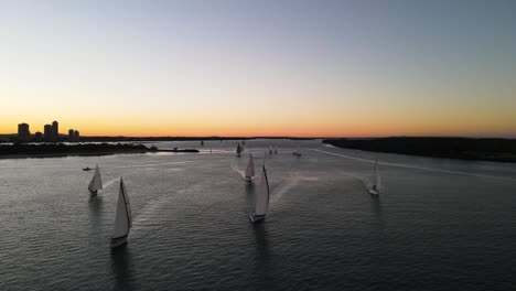 a group of moving sailing boats racing along the calm waters of a city harbor against a colorful sunset backdrop