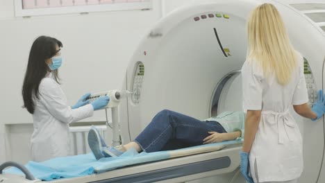 la paciente está siendo sometida a una tomografía computarizada o resonancia magnética bajo la supervisión de dos radiólogos calificados con máscara y guantes en una clínica médica moderna. la paciente está acostada en una mesa de tomografías computarizadas o mri