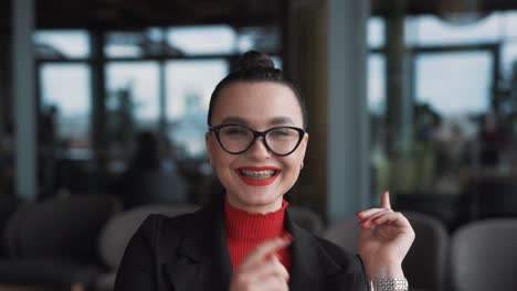 El-Retrato-Muestra-A-Una-Hermosa-Joven-Con-Gafas-Y-Aparatos-Ortopédicos,-Una-Profesional-Independiente,-Sentada-En-Un-Restaurante-Elegante,-Vestida-Con-Traje-De-Negocios,-Sonriendo-Y-Señalando-Sus-Aparatos-Ortopédicos.