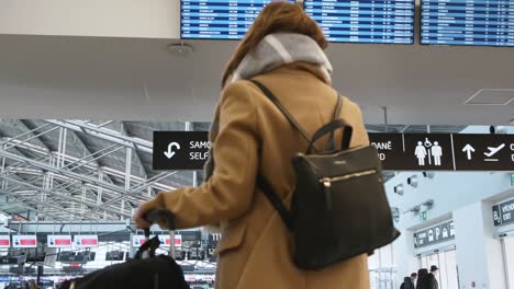 slightly blurred rear view female passenger with black bag on her back, standing in front of signs, looking at arrival and departure board at prague airport, czech republic. low season travel concept.