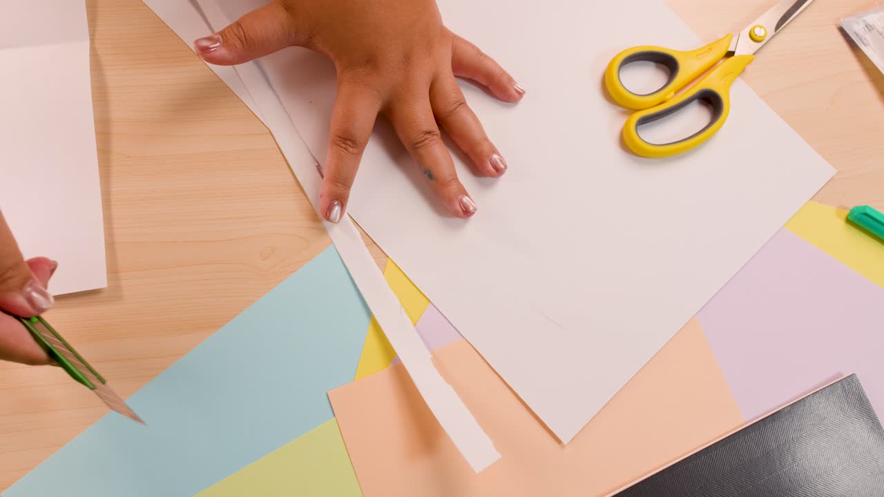 Hand uses green craft knife to cut white paper strip on pastel sheets, overhead view