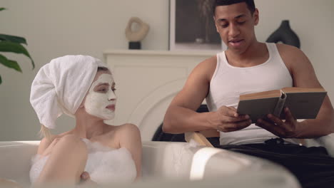 female with face mask taking a relaxing bath, black male reads a book