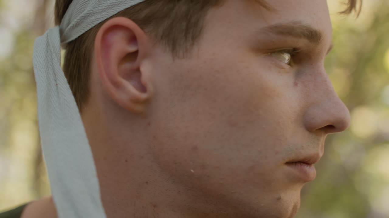 Close up view of soldier wearing green headband pulling strap over forehead with focused gaze, freckles and stubble visible on skin, blurred woodland background, intense yet calm expression