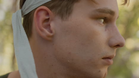 close up view of soldier wearing green headband pulling strap over forehead with focused gaze, freckles and stubble visible on skin, blurred woodland background, intense yet calm expression