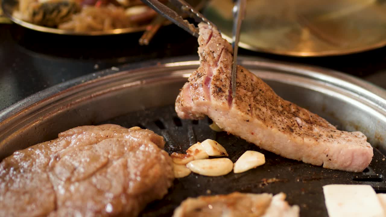 Hand uses tongs and scissors to slice pork belly on tabletop grill, warm indoor lighting
