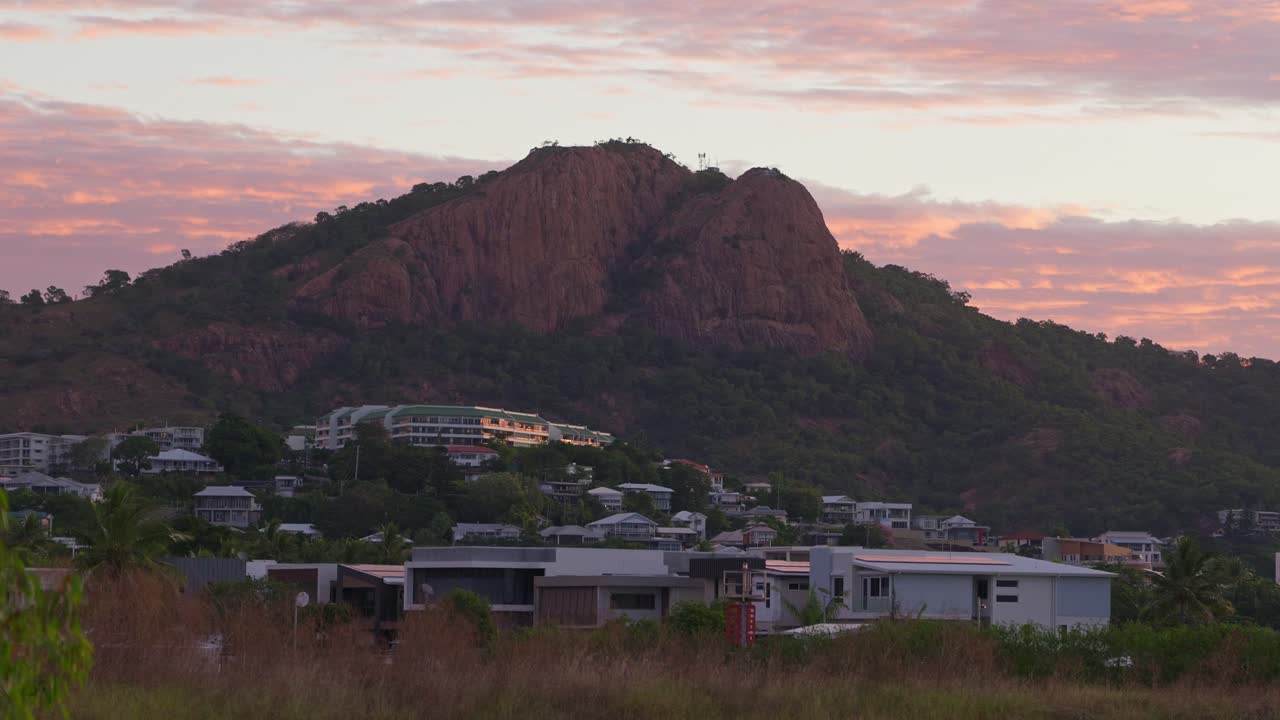 Castle Hill in Townsville at sunrise, peaking from behind grass blades with houses glowing in the pink light at the base