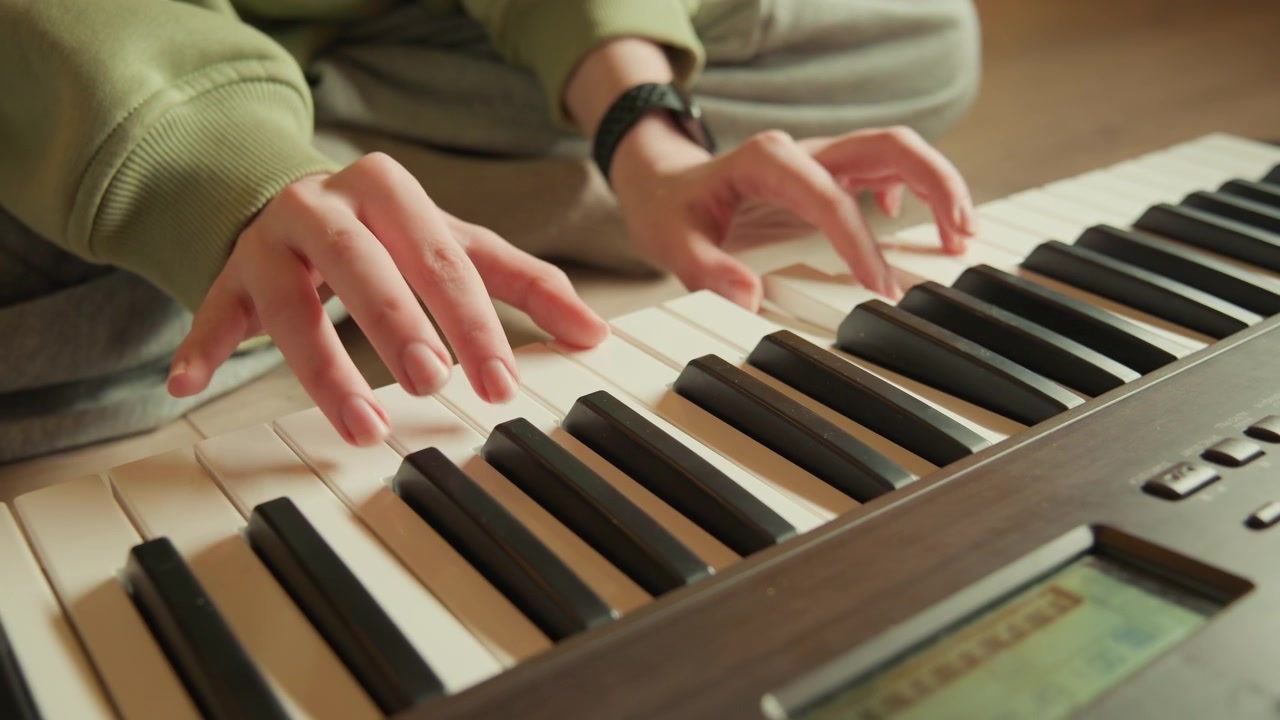 Voksne hænder i grønne lange ærmer og armbåndsur spiller på et elektronisk keyboard fra lav vinkel, fingre trykker på blanke sorte og hvide tangenter med jævn rytme, blødt lys og tydelig bevægelse.